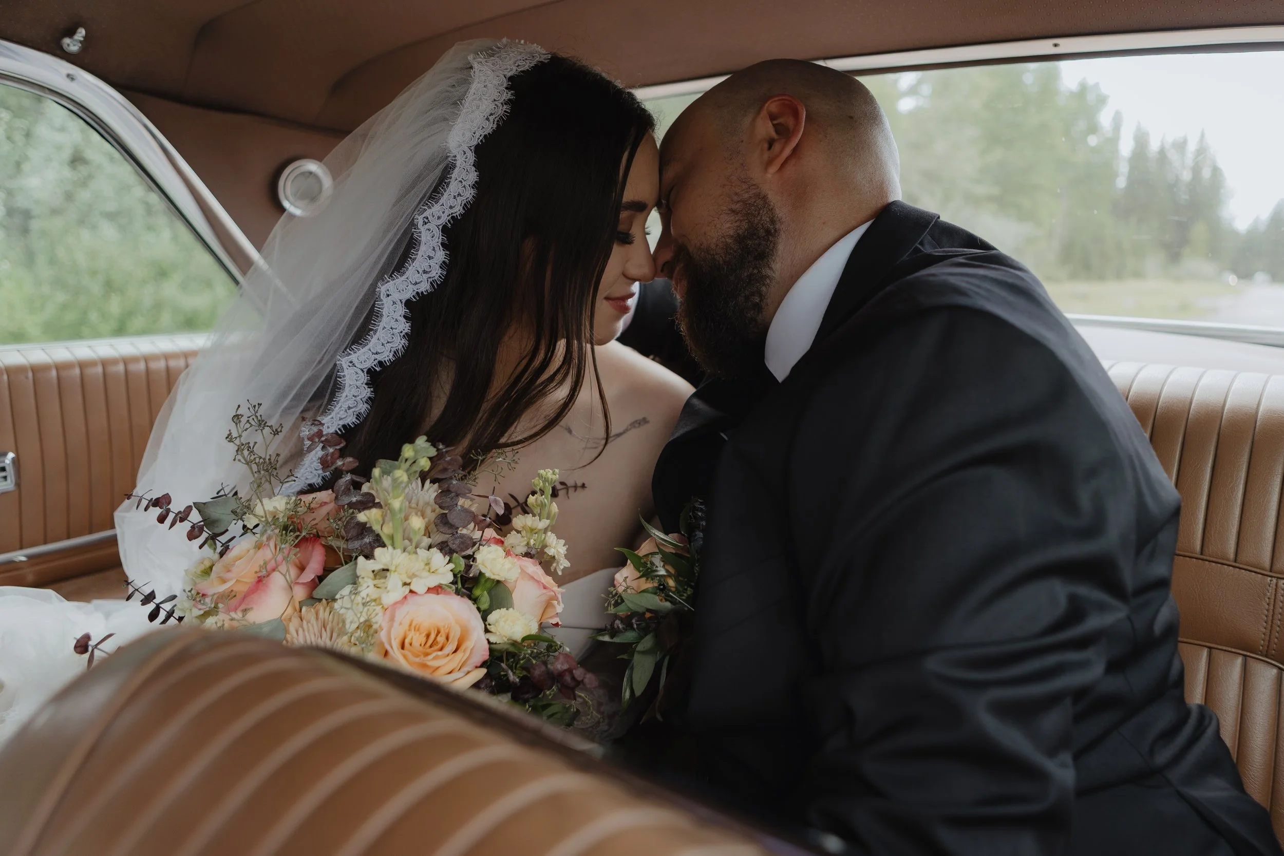 A bride and groom sharing a tender moment inside a vintage car, with their foreheads touching and eyes closed. The bride is wearing a lace-edged veil and holding a bouquet of flowers, while the groom is dressed in a black tuxedo. The background shows blurred trees and greenery outside the car window.