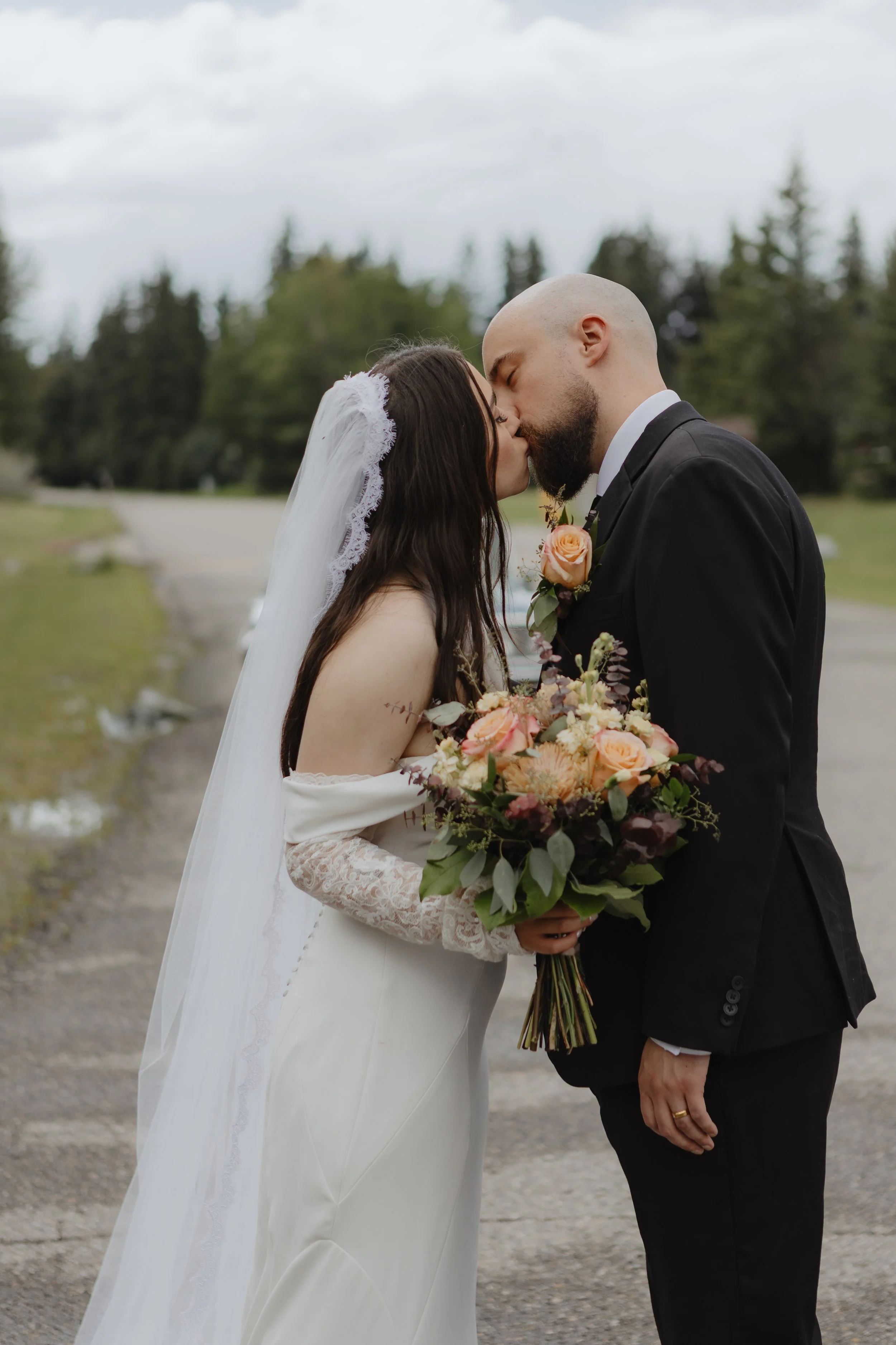 A bride and groom sharing a kiss outdoors, with the bride holding a bouquet of flowers and wearing a white wedding dress and veil, and the groom dressed in a black suit with a boutonnière.