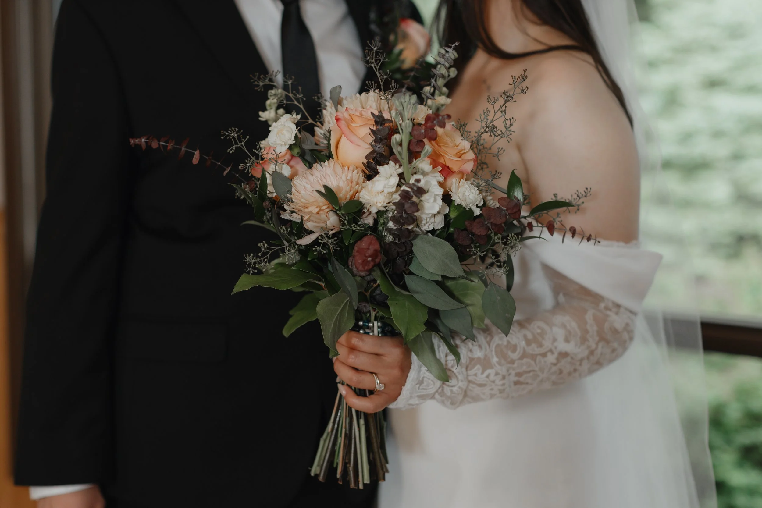 A bride and groom holding a bouquet of flowers on their wedding day. The bride is wearing a lace wedding dress with an off-shoulder design, and the groom is dressed in a black tuxedo. The bouquet includes peach, white, and dark purple flowers with greenery.