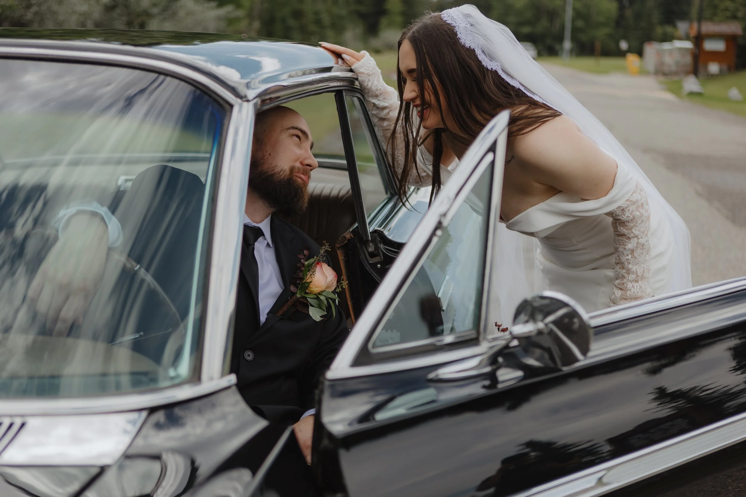Bride leaning into car window speaking to groom, both smiling, outdoors on a rural road.