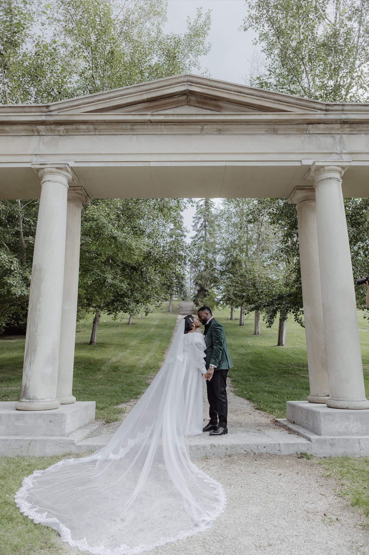 A bride and groom holding hands and sharing a moment underneath a classical stone gazebo in a green park.