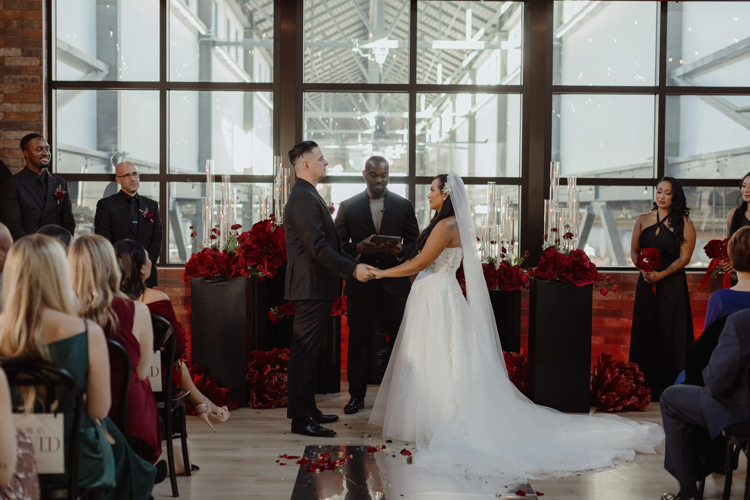 A wedding ceremony with a bride and groom holding hands, standing in front of an officiant, with bridesmaids and groomsmen in the background, inside a venue with large windows and red floral decorations.