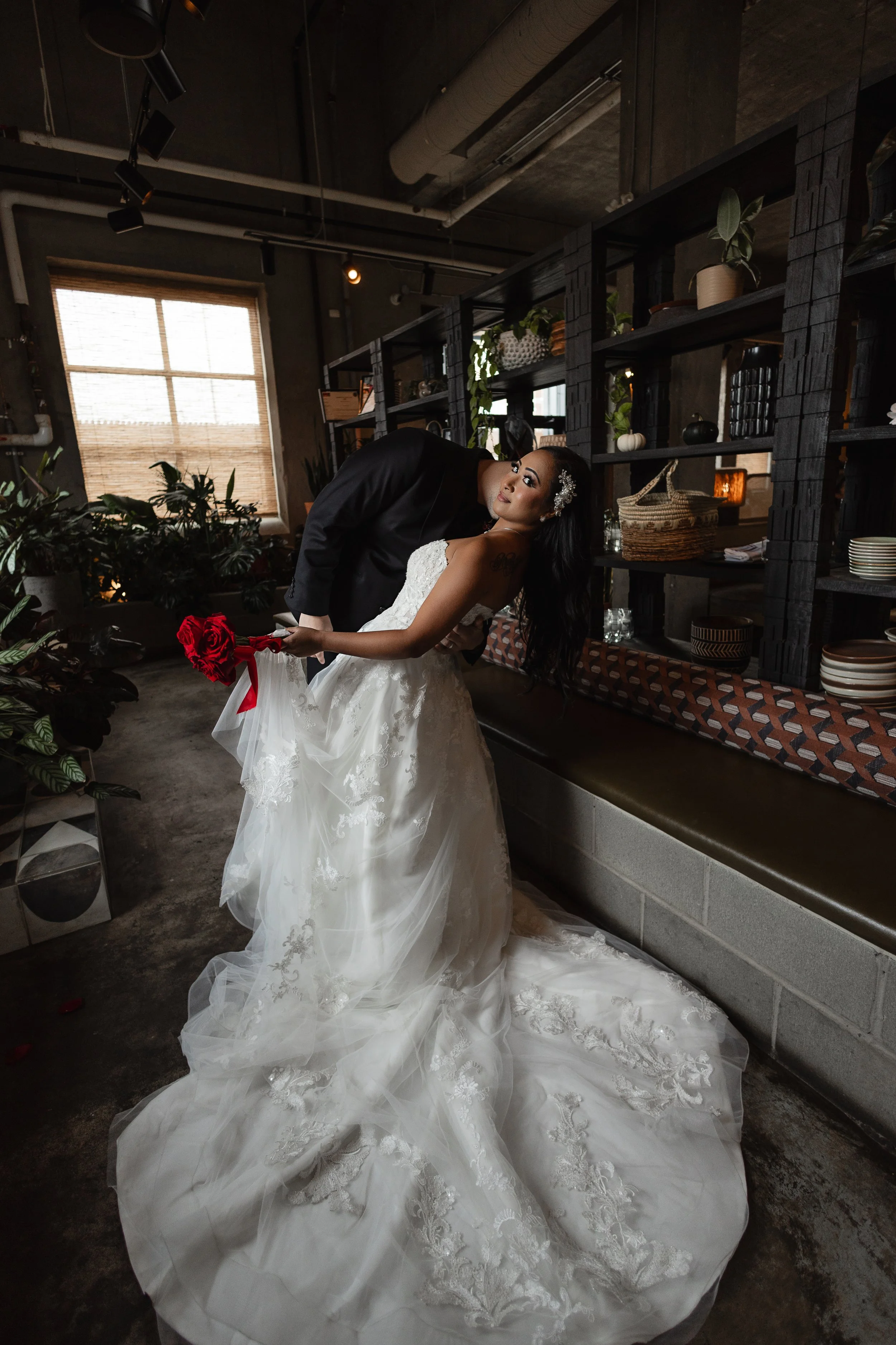 A bride in a white wedding gown holding a red rose, leaning back as a groom in a black suit dips her, in an industrial-style room with dark shelves, plants, and a large window with blinds.