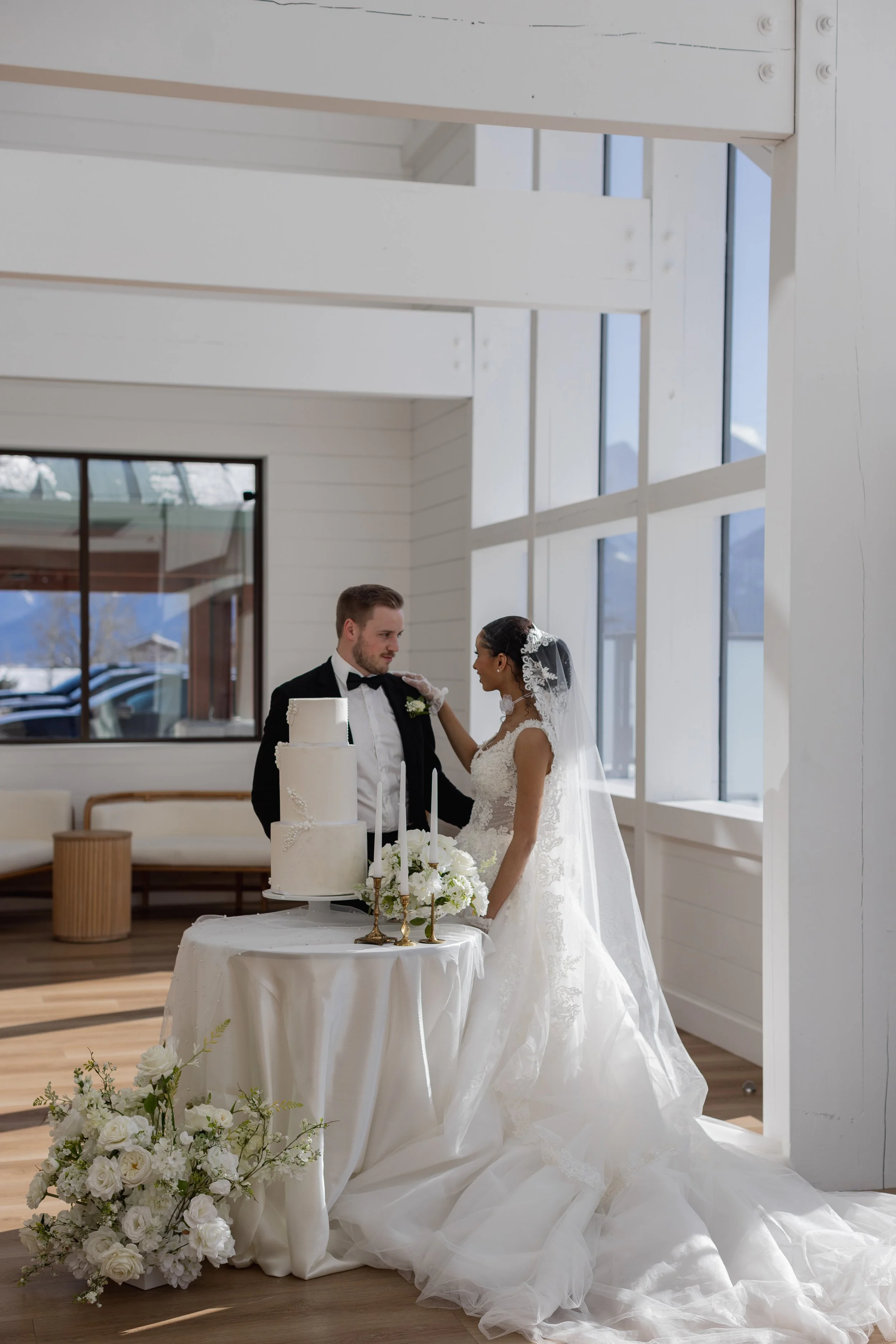 A bride and groom at their wedding, standing beside a table with a wedding cake and floral arrangements, inside a bright, modern venue with large windows.