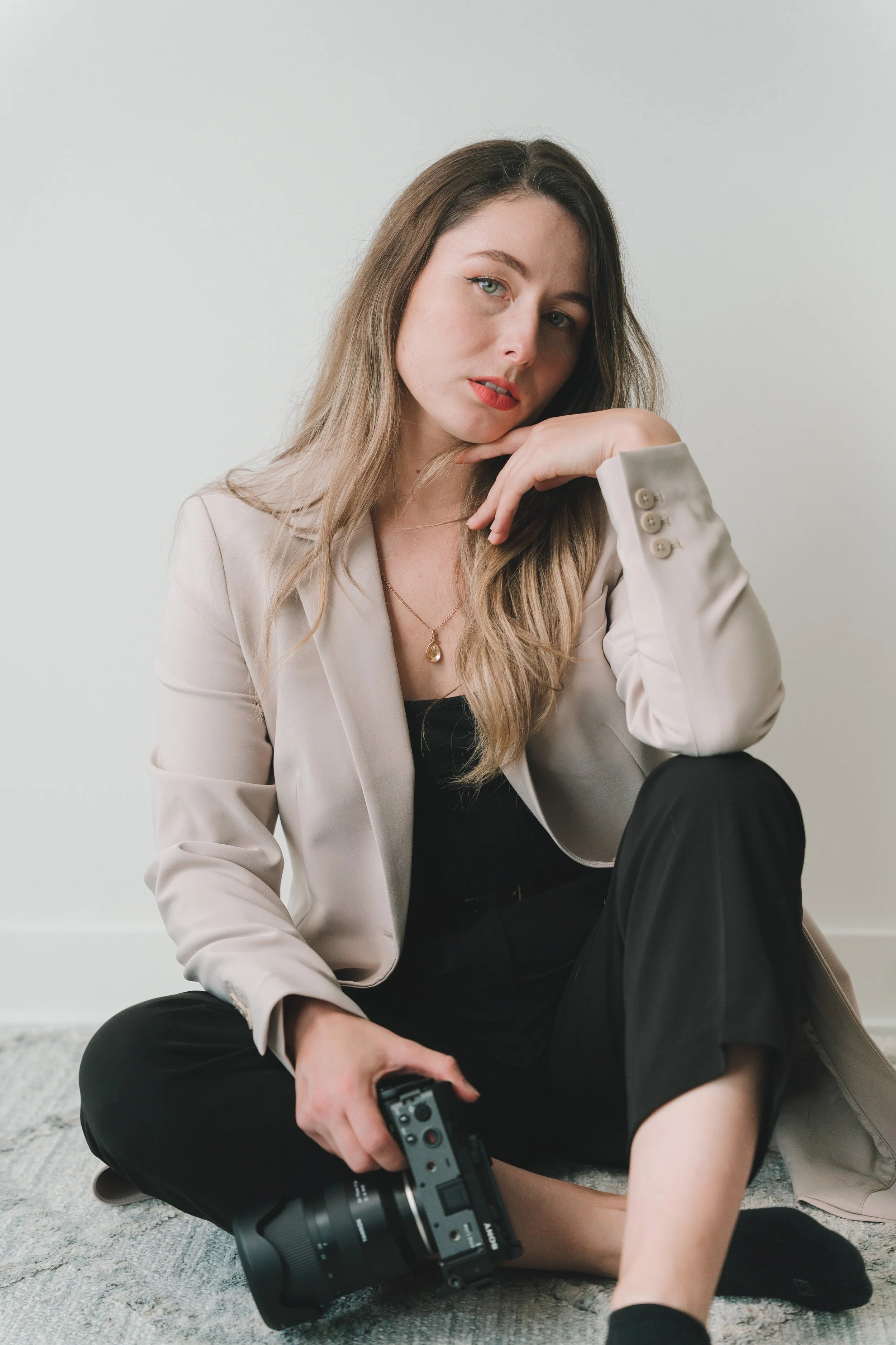 A young woman with long, wavy blonde hair sitting on a carpeted floor, holding a camera in her right hand, wearing black pants, a beige blazer, and a black top, with a neutral expression, against a plain light-colored background.