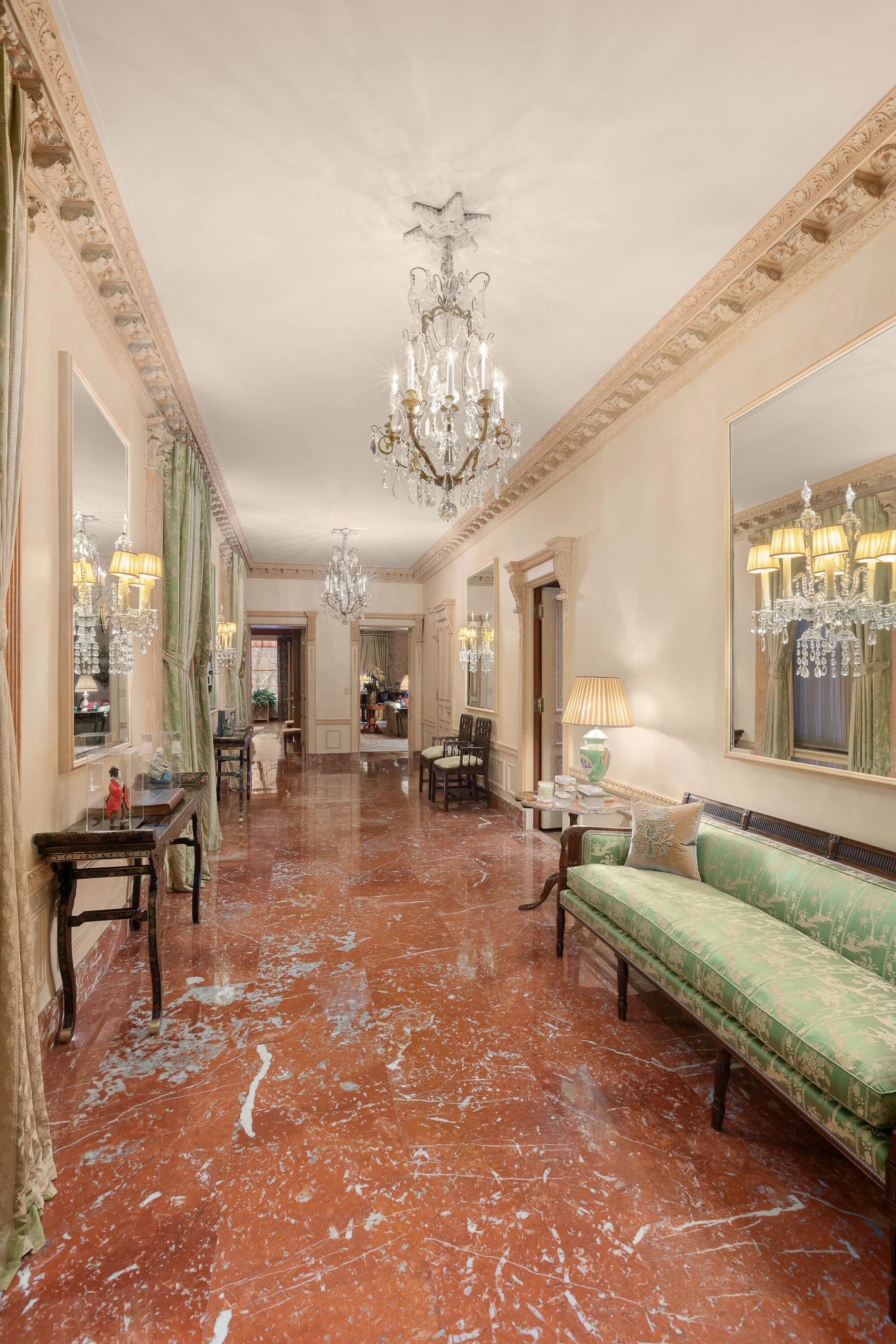 Elegant hotel hallway decorated with chandeliers, large mirrors, green velvet upholstery, and marble flooring.
