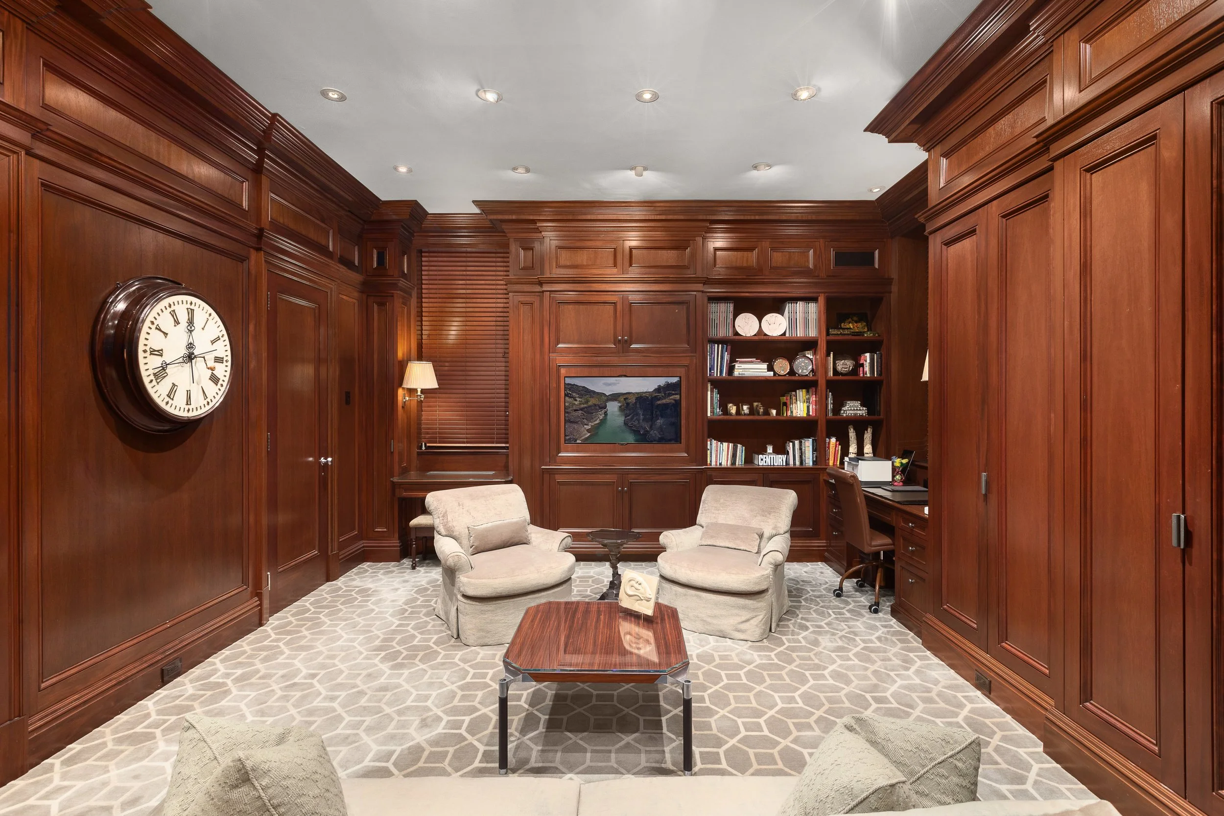 A cozy home office with wood-paneled walls, a patterned area rug, two beige armchairs, a small wooden coffee table, built-in wooden bookshelves filled with books and decor items, a wall-mounted television, a desk with a chair, and a large clock on the wall.