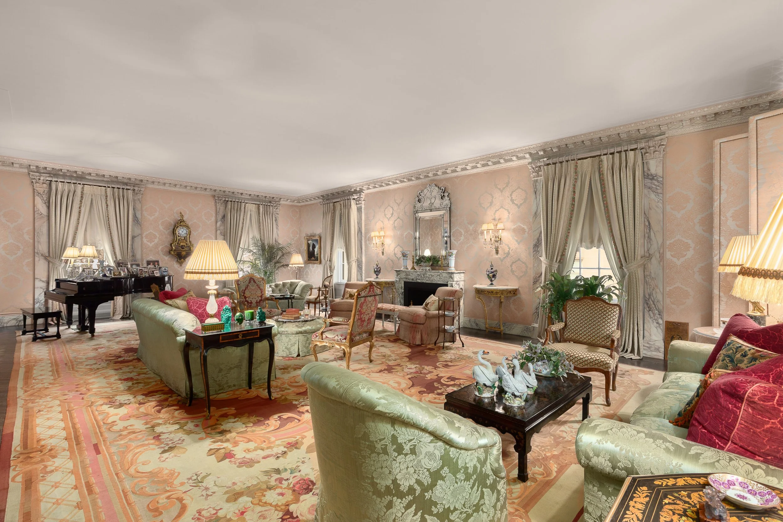 Elegant living room with pastel-colored furniture, a fireplace with a mirror above it, floral rugs, vintage lamps, and large windows with draped curtains.