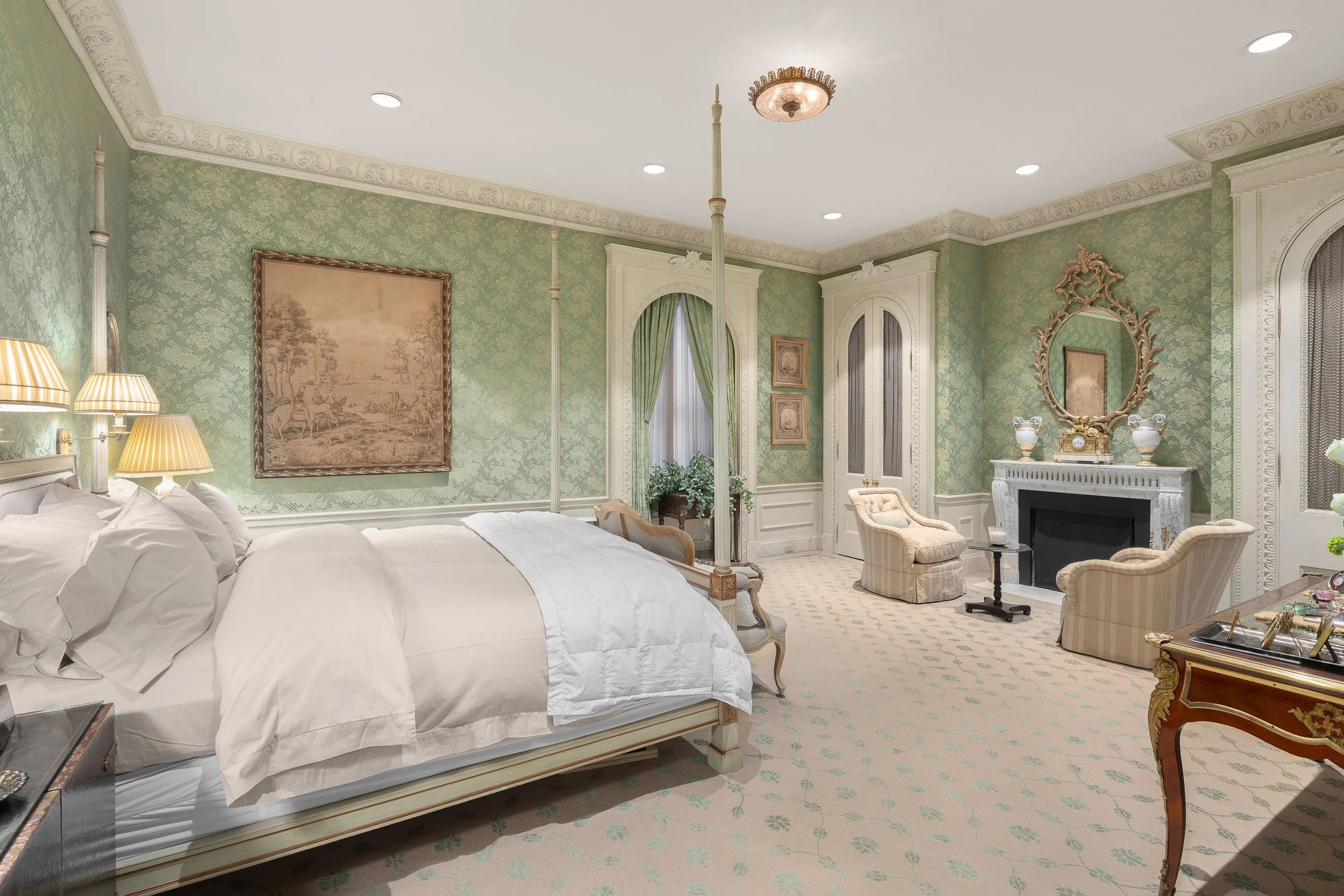A luxurious bedroom with green wallpaper, white ornate crown molding, and a four-poster bed with white linens. There are lamps beside the bed, a decorated fireplace with an ornate mirror above, and elegant chairs near the fireplace. The room also has a window with green curtains, artwork on the walls, and a decorative carpet.