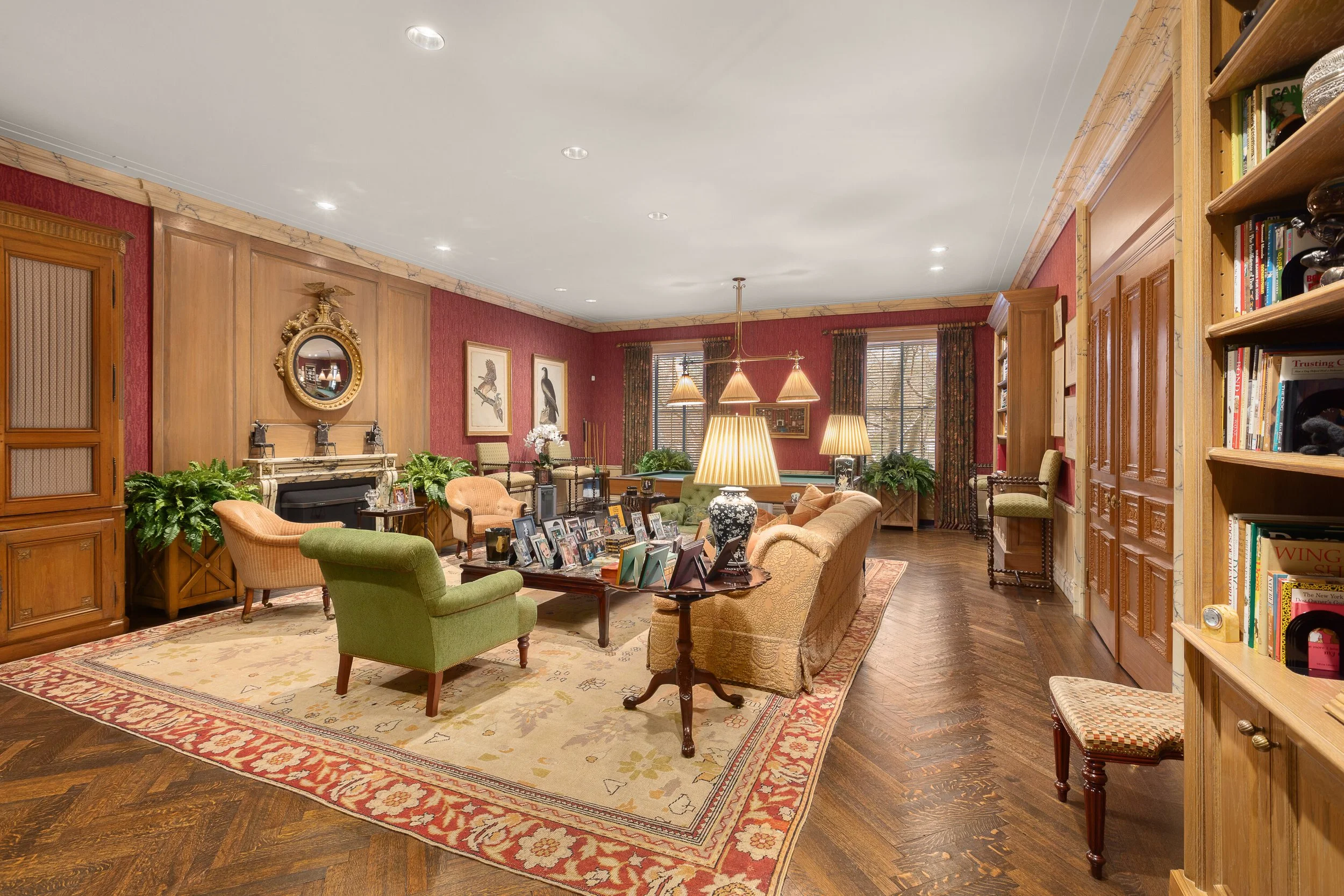 A spacious, elegant living room with red walls, wooden accents, and festive decorations. Multiple sofas, armchairs, and lamps are arranged around a central coffee table with framed photos. There are large windows with floral curtains and a bookshelf on the right. The hardwood floor is partially covered by a large area rug.