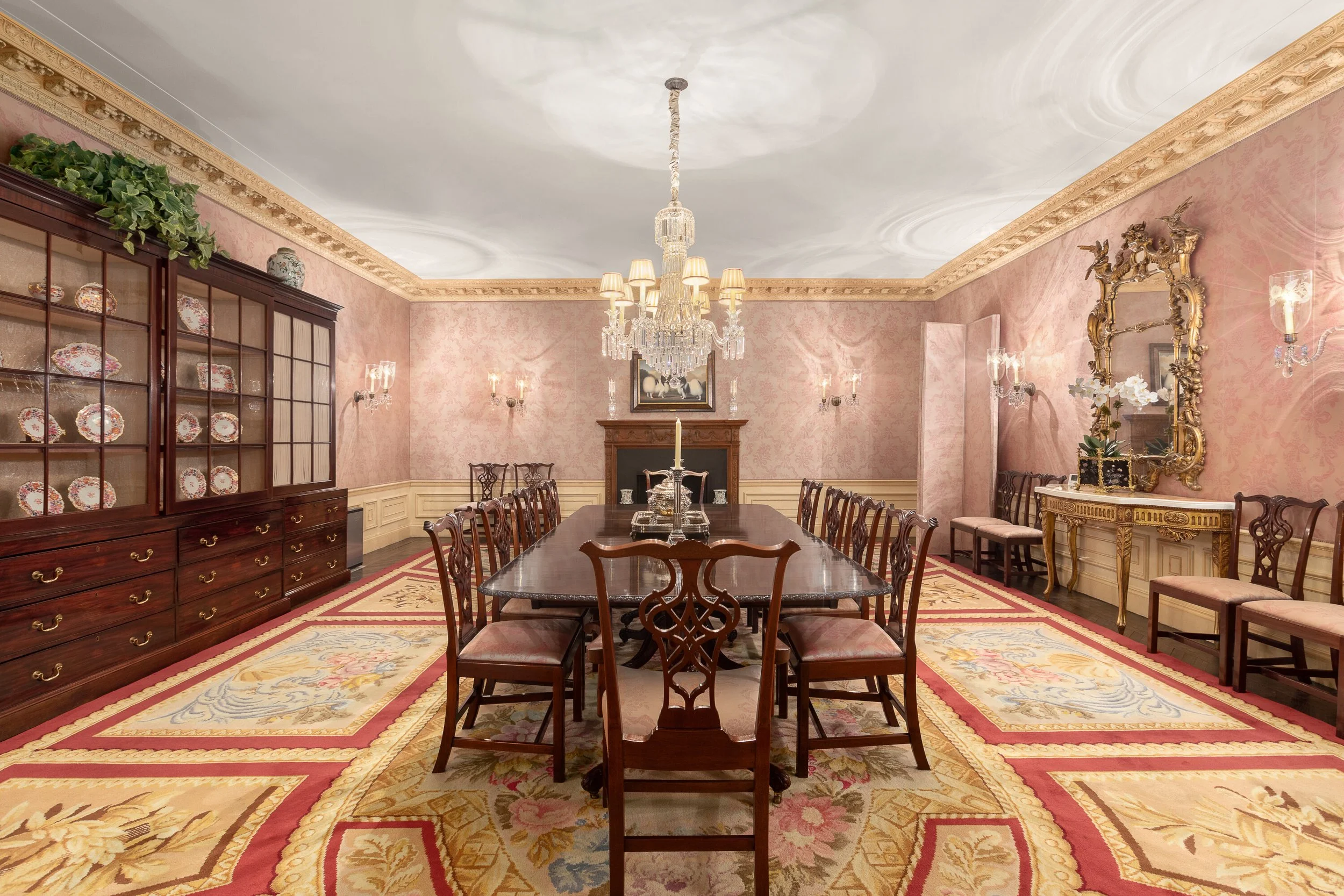 Elegant dining room with a long wooden table, pink wallpaper, a chandelier, a fireplace, and ornate furniture.