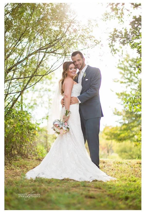 A bride and groom standing outdoors, embracing each other. The bride is in a white wedding gown holding a bouquet, and the groom is in a dark suit. They are smiling and surrounded by trees and greenery.