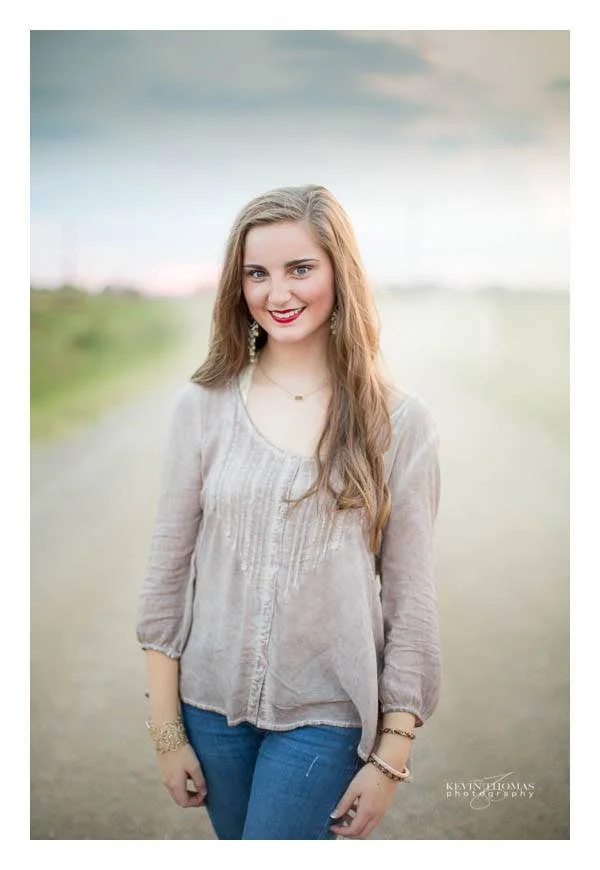 A young woman with long, wavy light brown hair standing outdoors in a blurred natural setting with green grass and a cloudy sky, smiling at the camera, wearing a light-colored blouse and blue jeans.