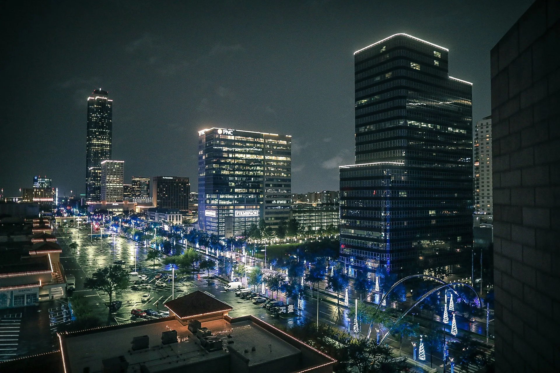 Nighttime cityscape of downtown with illuminated skyscrapers, decorated streets, and a parking lot.