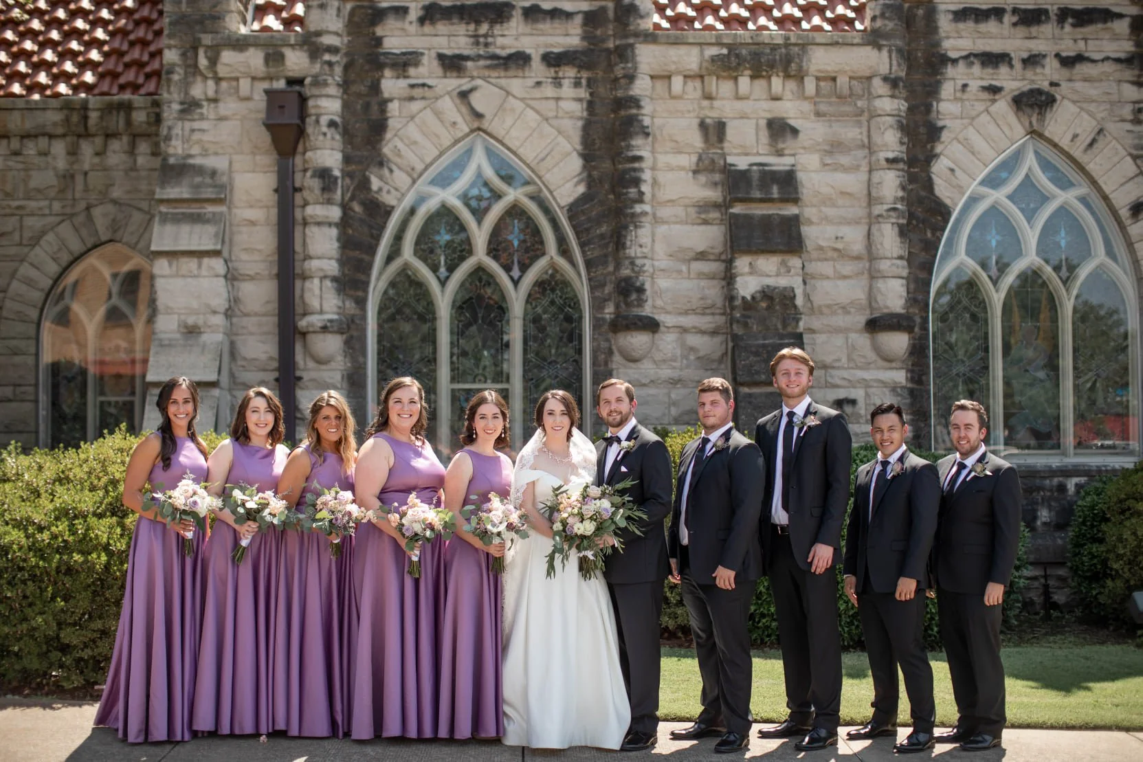 Wedding party in front of a stone church, with six bridesmaids in purple dresses holding bouquets, two bridesmaids in white, and five groomsmen in dark suits with ties, standing on a grassy lawn.