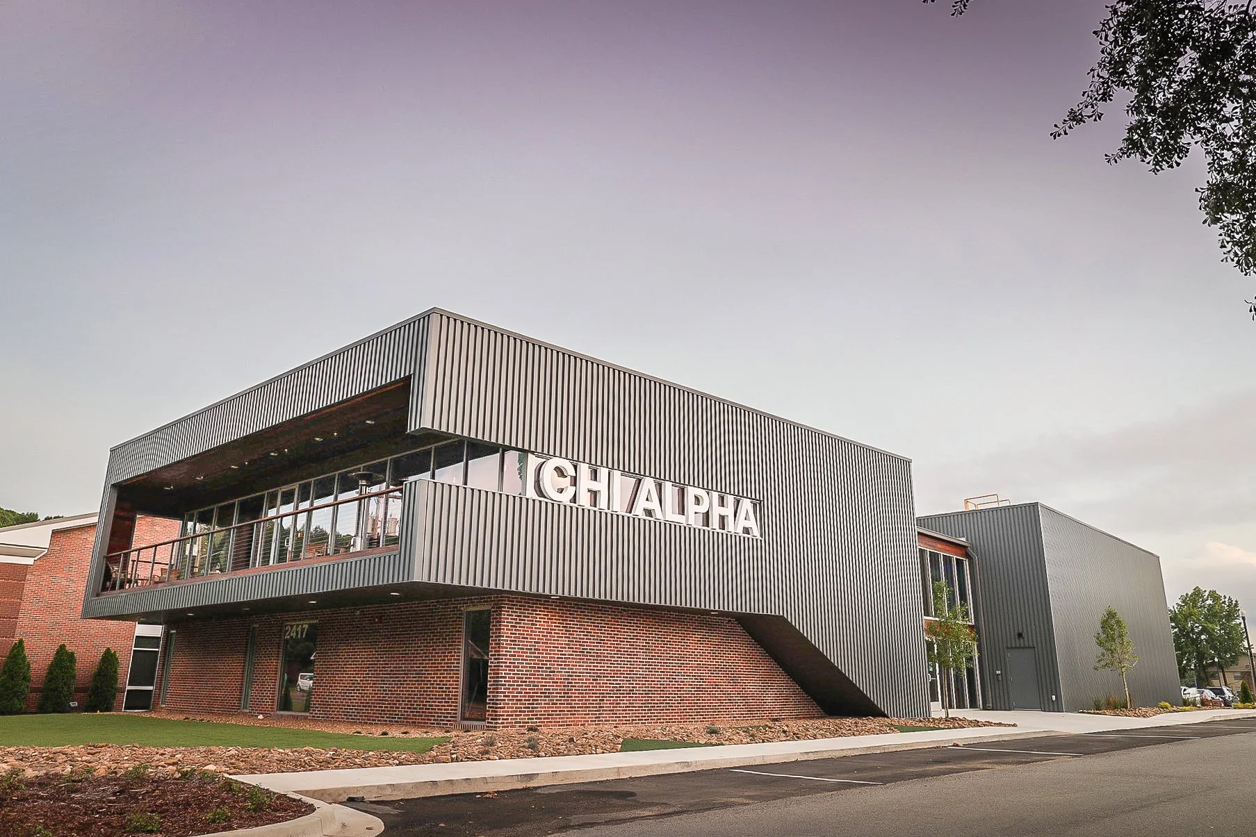 Modern commercial building with metal siding and brick base, featuring large windows and surrounded by a parking lot and landscaping with small trees and bushes.