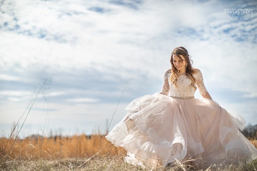 A woman in a white wedding dress sitting outdoors in a field with tall grasses, smiling, with partly cloudy skies overhead.