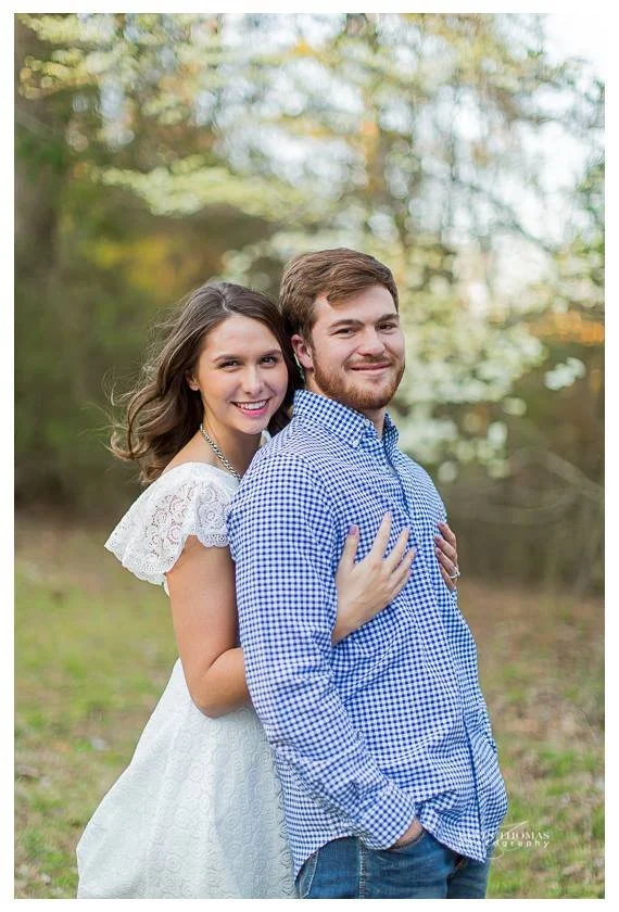 A young woman in a white dress and a young man in a blue checkered shirt standing outdoors in a wooded area during daytime, smiling and embracing each other.
