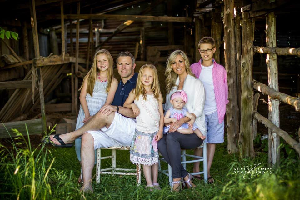 Family portrait of six people with three children, sitting and standing in front of a rustic barn, outdoors with green grass and plants.