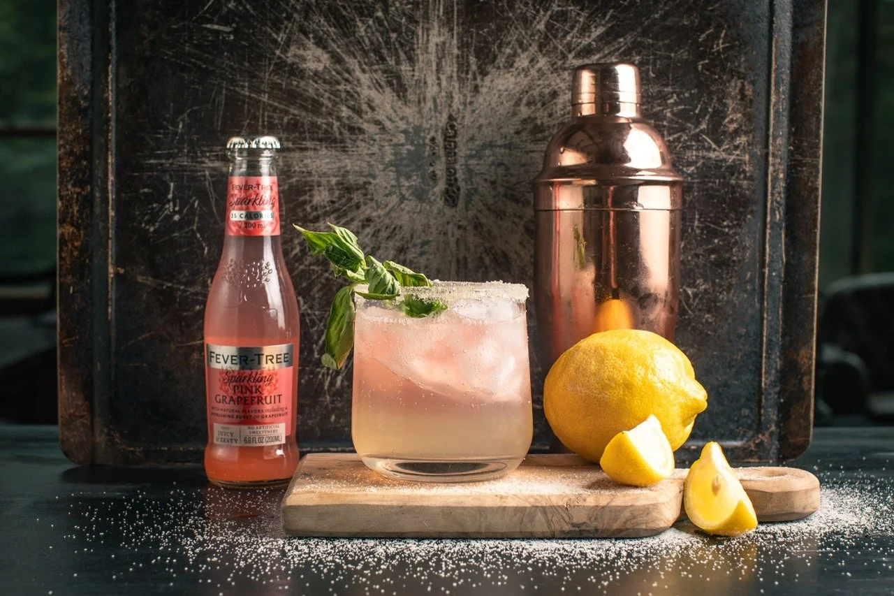 A cocktail glass with a pink drink garnished with a basil leaf, placed on a wooden cutting board. To the left, a bottle of Fever-Tree sparkling pink grapefruit soda. To the right, a whole lemon with lemon wedges, a copper cocktail shaker, and a rusti