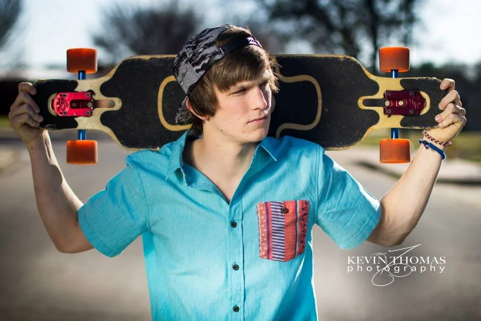 A young man wearing a black and gray camouflage baseball cap and a bright blue shirt with a colorful pocket, holding a skateboard on his shoulders at an outdoor skate park.
