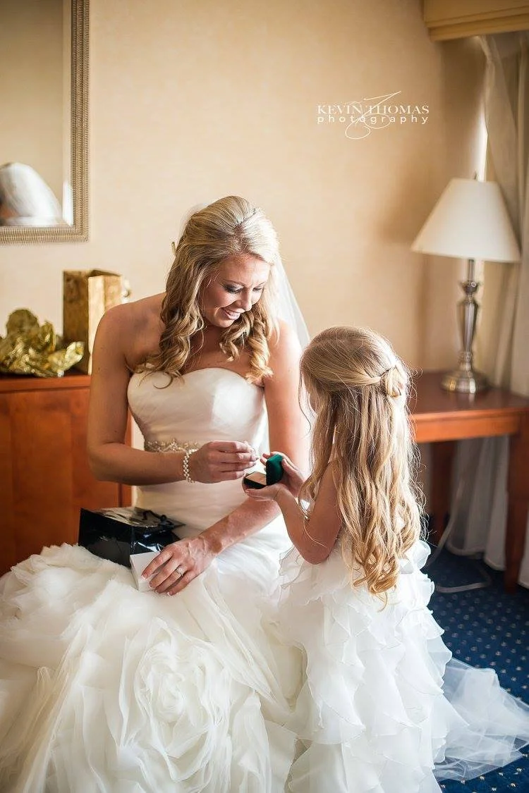 A bride in a white wedding gown smiling at a young girl with long blonde hair, who is also dressed in a white dress, as they engage in a gift exchange in a warmly lit room.