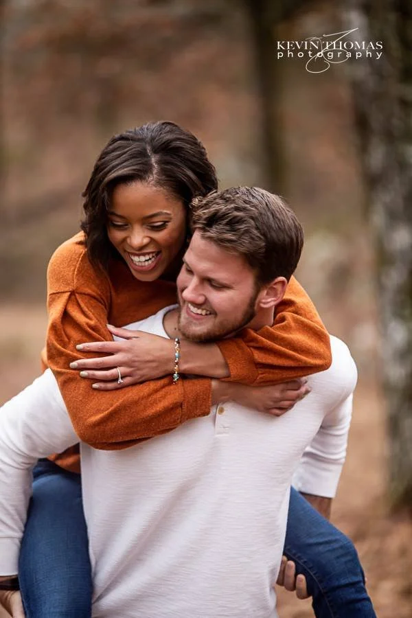 A smiling couple outdoors in fall, with the woman on the man's back, both enjoying each other's company.