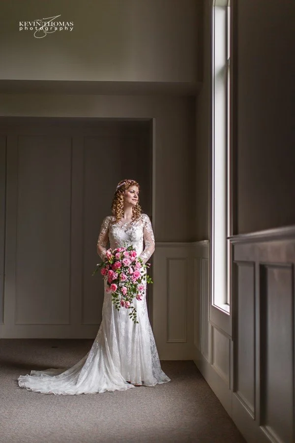 Bride in a long-sleeved lace wedding gown holding a cascading bouquet of pink roses and greenery, standing in a softly lit room near a large window, with a serene expression.