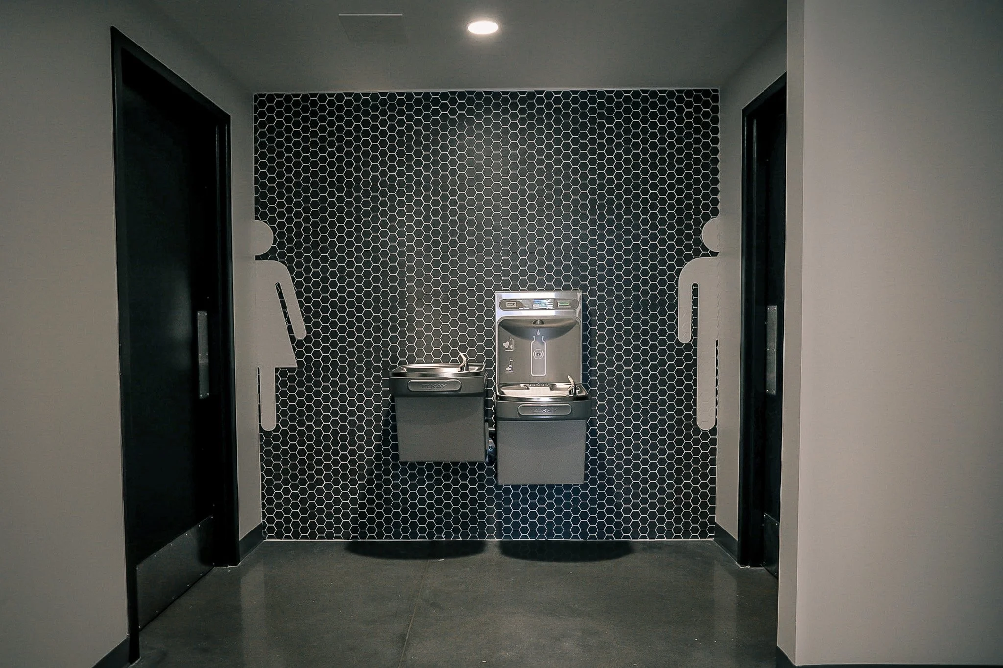 Public restroom with two sinks and black hexagonal tile wall, woman and man symbols on either side.
