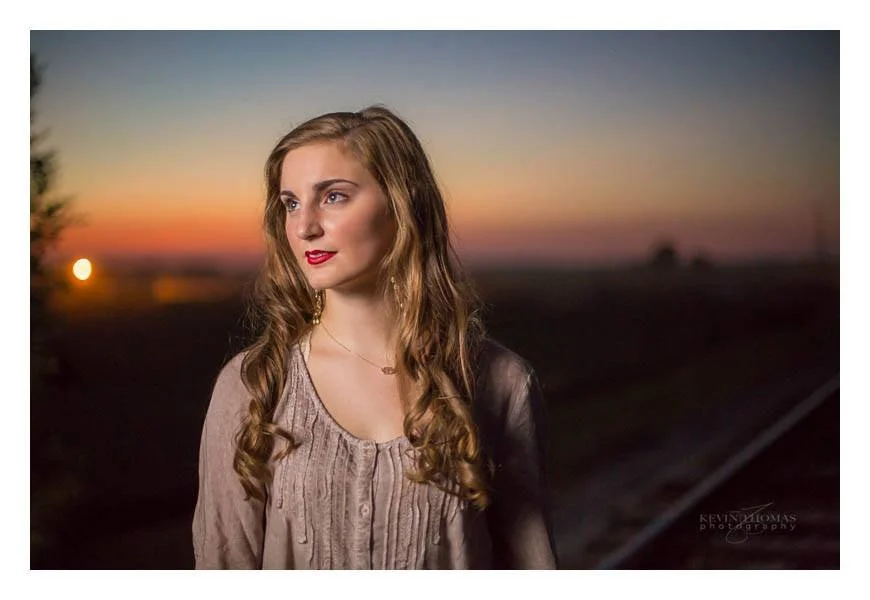 A woman with long wavy hair and red lipstick, standing outdoors at sunset, wearing a beige blouse with pleats and a black leather jacket.