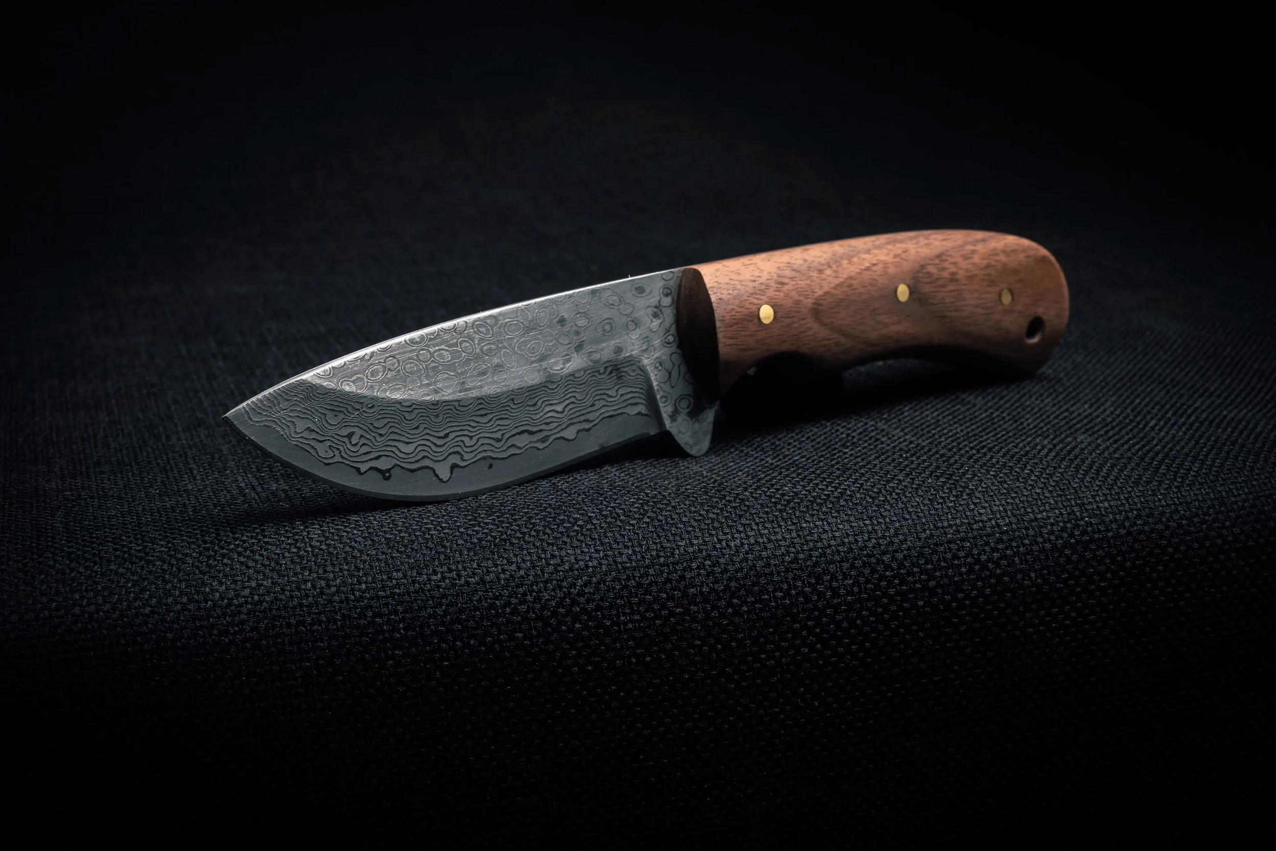 A handcrafted knife with a Damascus steel blade and a wooden handle, resting on a black fabric surface.