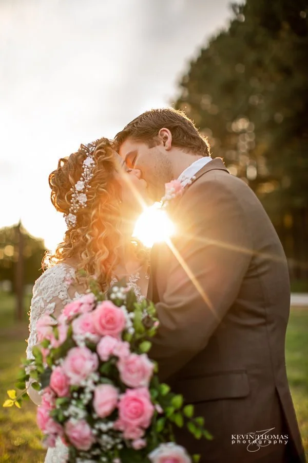 A bride and groom kissing outdoors at sunset, with the sun shining behind them and a bouquet of pink roses in the foreground.
