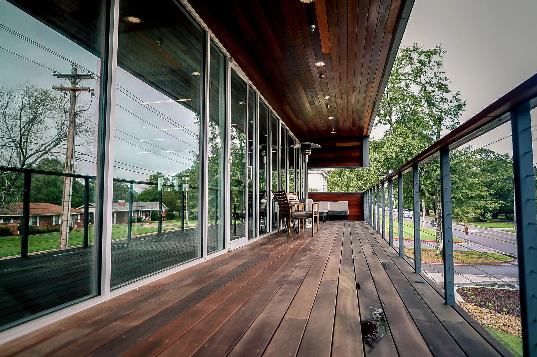 Empty balcony with wooden flooring, glass sliding doors, and outdoor furniture, overlooking a suburban street with trees.