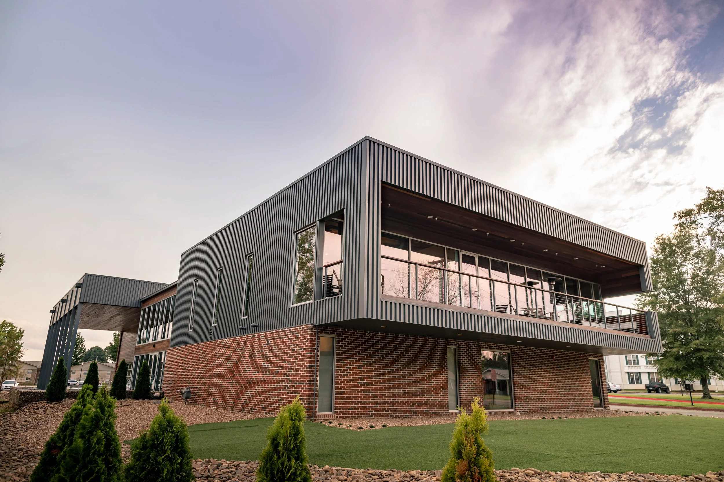 Modern two-story building with metal siding on the upper level and brick on the lower level, large windows, and a balcony, surrounded by landscaped grass and small trees.