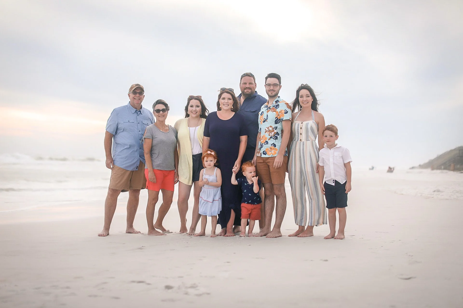 Family of eleven standing on the beach during sunset, smiling at the camera, with ocean waves in the background.