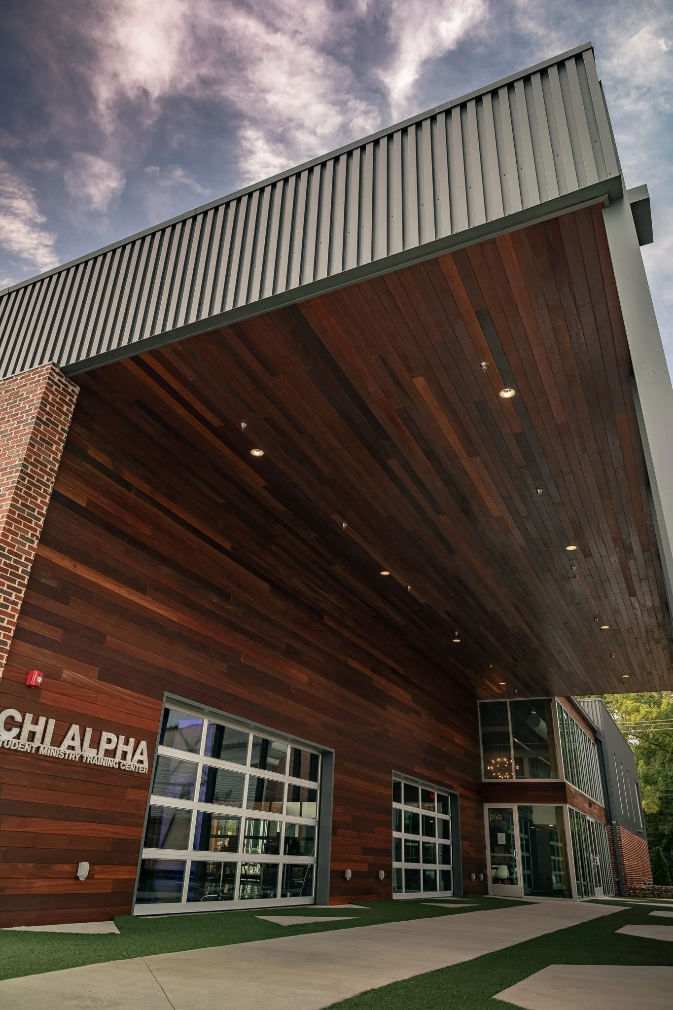 Front view of a modern building with a wooden and metal exterior, large glass windows, and a sign that reads 'CHIALPHA STUDENT MINISTRY TRAINING CENTER' on a sunny day.