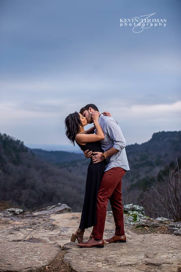A couple embraces and kisses on a rocky mountaintop with a scenic valley and hills in the background under an overcast sky.