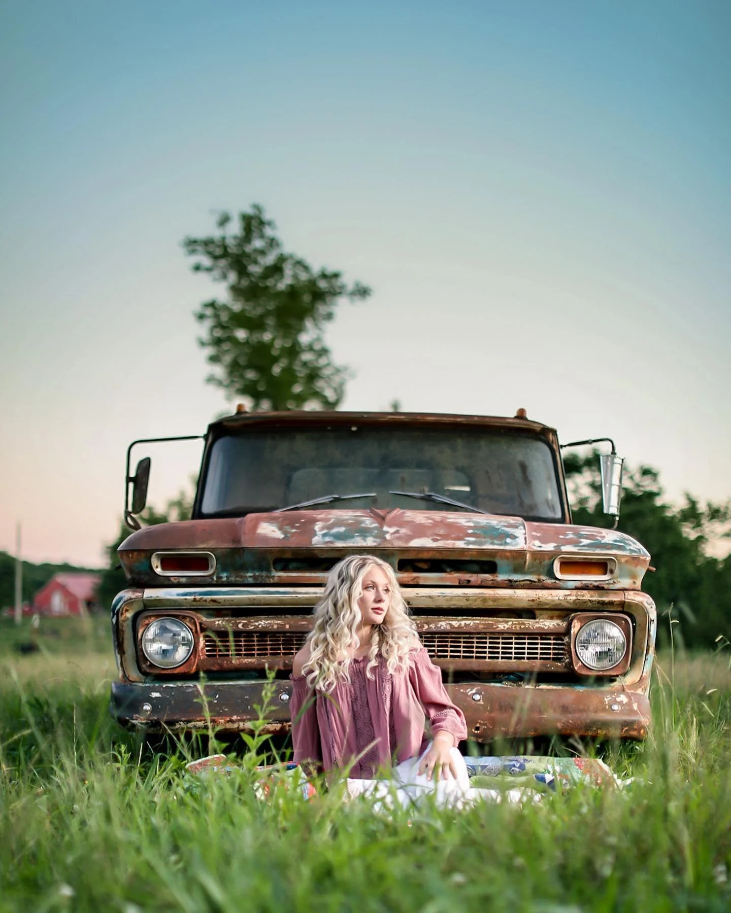 A woman with long, curly blonde hair wearing a pink off-shoulder dress, sitting in a grassy field in front of a rusted, old pickup truck during sunset.