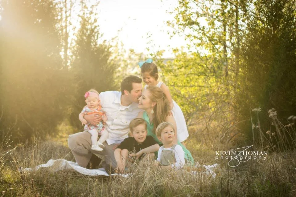 Family of six sitting on a blanket in a grassy field with trees in the background, smiling and enjoying outdoor time.