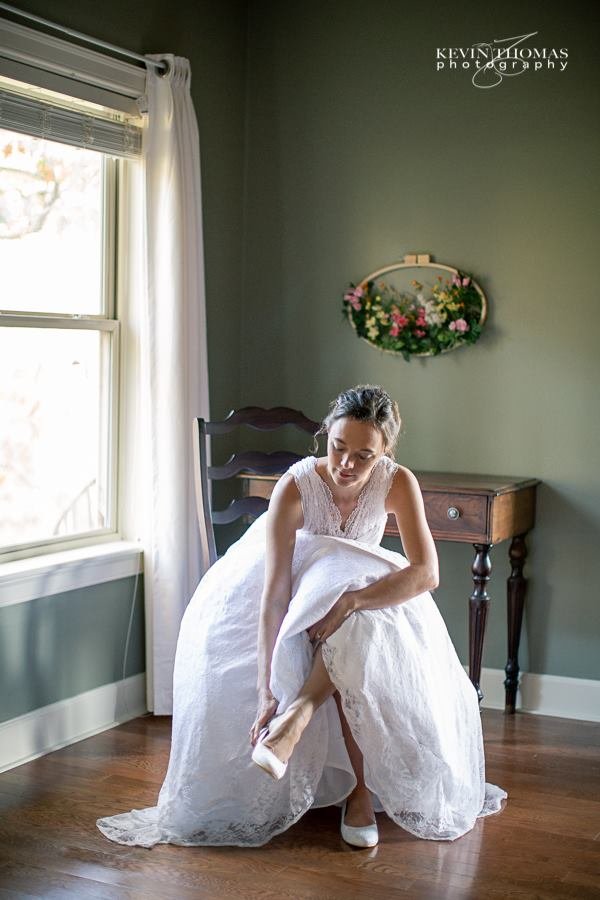 Bride in a white wedding dress adjusting her shoe while sitting on a wooden chair in a room with green walls and a large window.