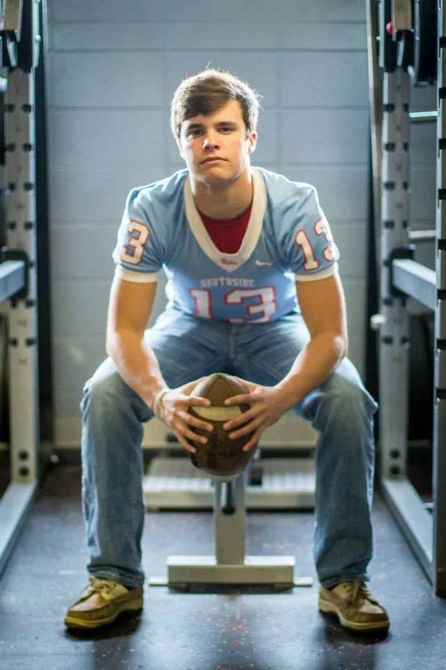 A young man in a blue sports jersey with the number 13, sitting on a bench, holding a football, inside a gym with gym equipment in the background.