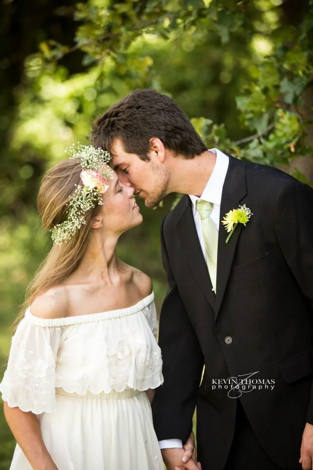 A couple, dressed in wedding attire, touching foreheads and holding hands outdoors on a green, leafy background.