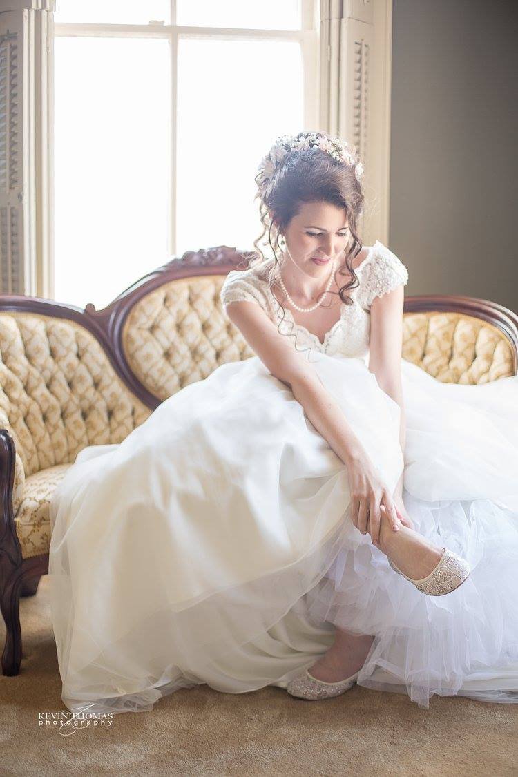Bride in a white wedding dress adjusting her shoe on a vintage sofa beside a window.