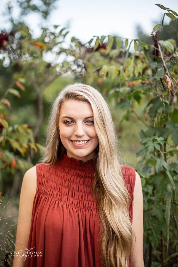 A young woman with long blonde hair smiling outdoors, wearing a sleeveless, red, gathered-top dress, surrounded by green foliage.