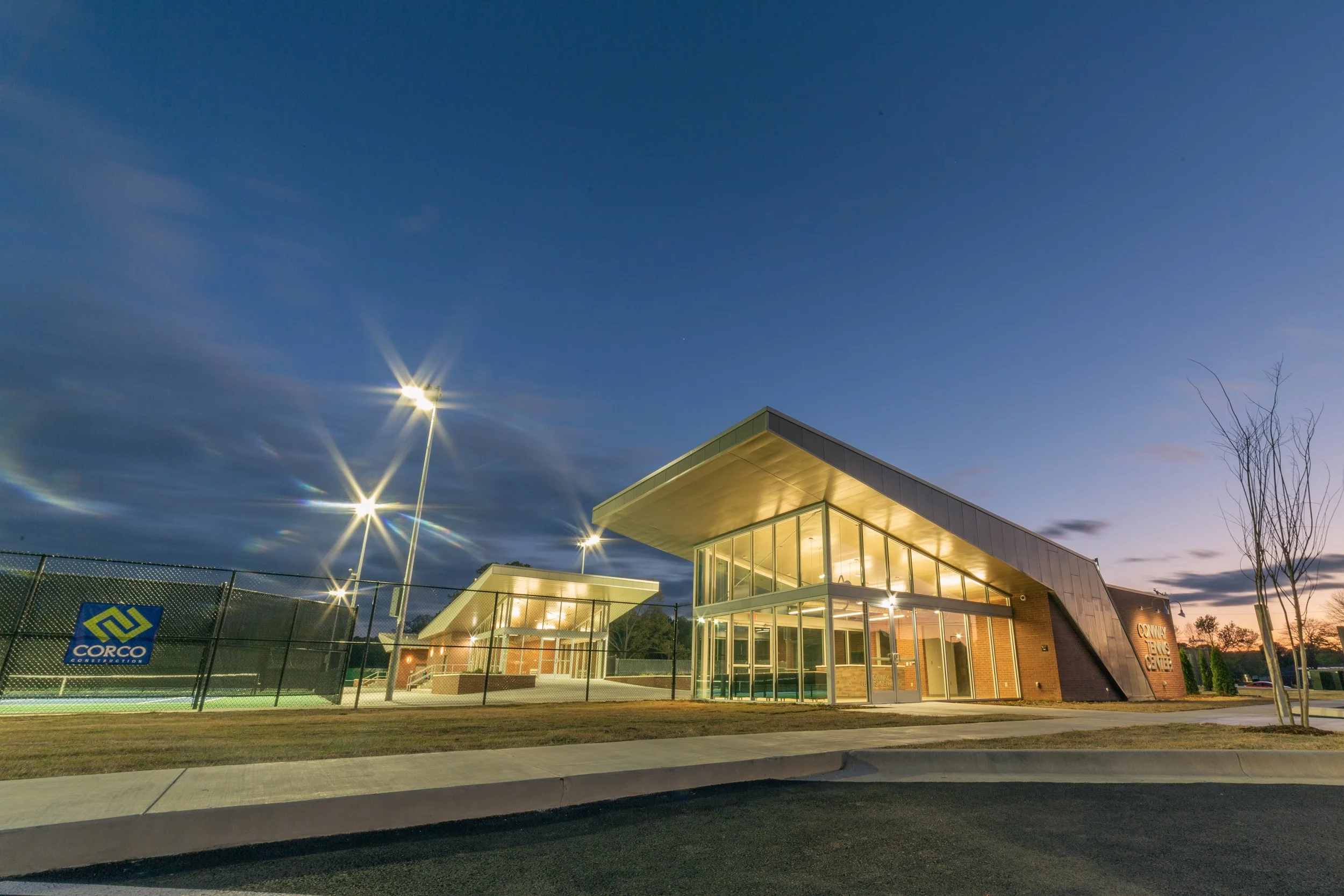 Exterior view of a modern sports complex at dusk with illuminated indoor tennis courts, a glass building, and a tennis court with high fencing, surrounded by a paved sidewalk and trees.