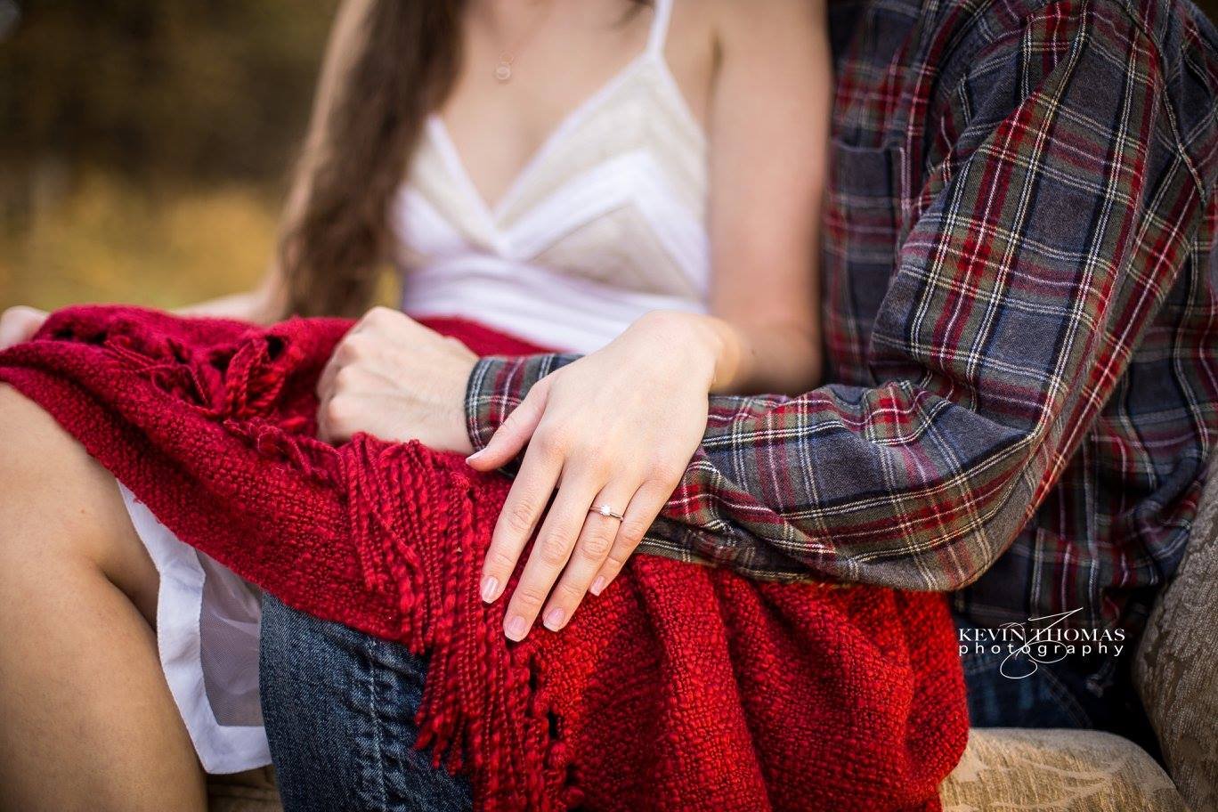 A woman and a man sitting close together outdoors, with the woman's hand resting on the man's knee showing an engagement ring, both wearing casual clothing.