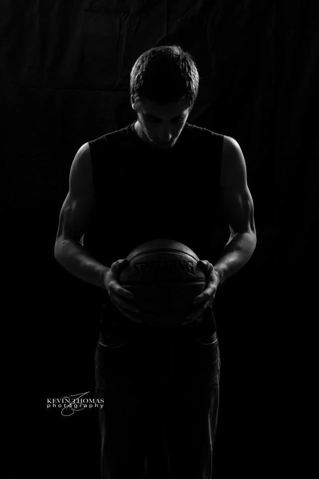Black and white photo of a young man holding a basketball with head bowed, in a dark setting with his upper arms visible.