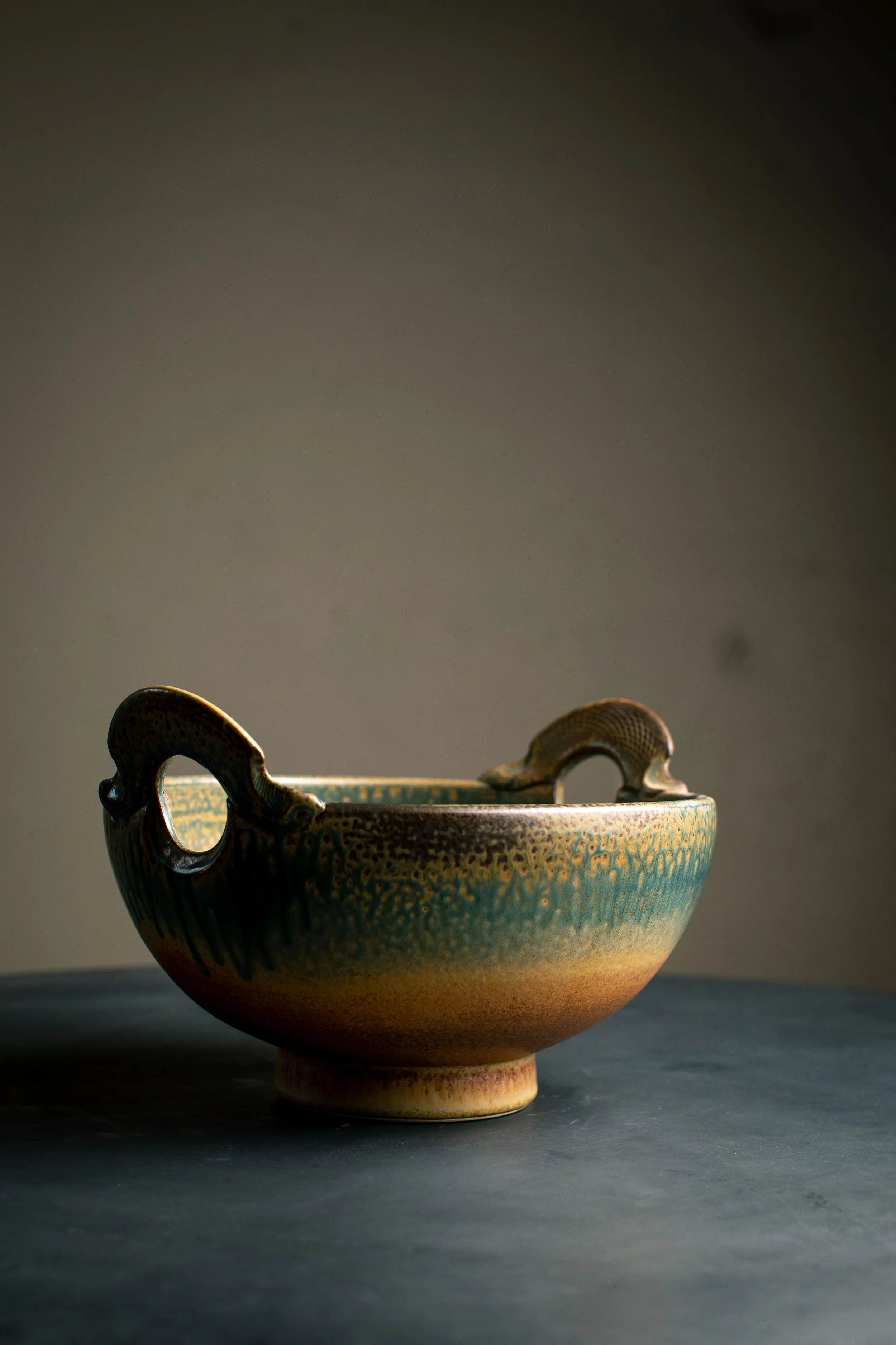 A ceramic bowl with two ornate handles sits on a dark surface against a plain, neutral background.