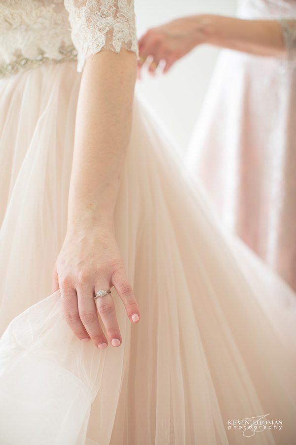 Close-up of a bride's hand with a wedding ring, holding her wedding gown's fabric. The gown has lace details on the bodice and a flowy, light-colored skirt.