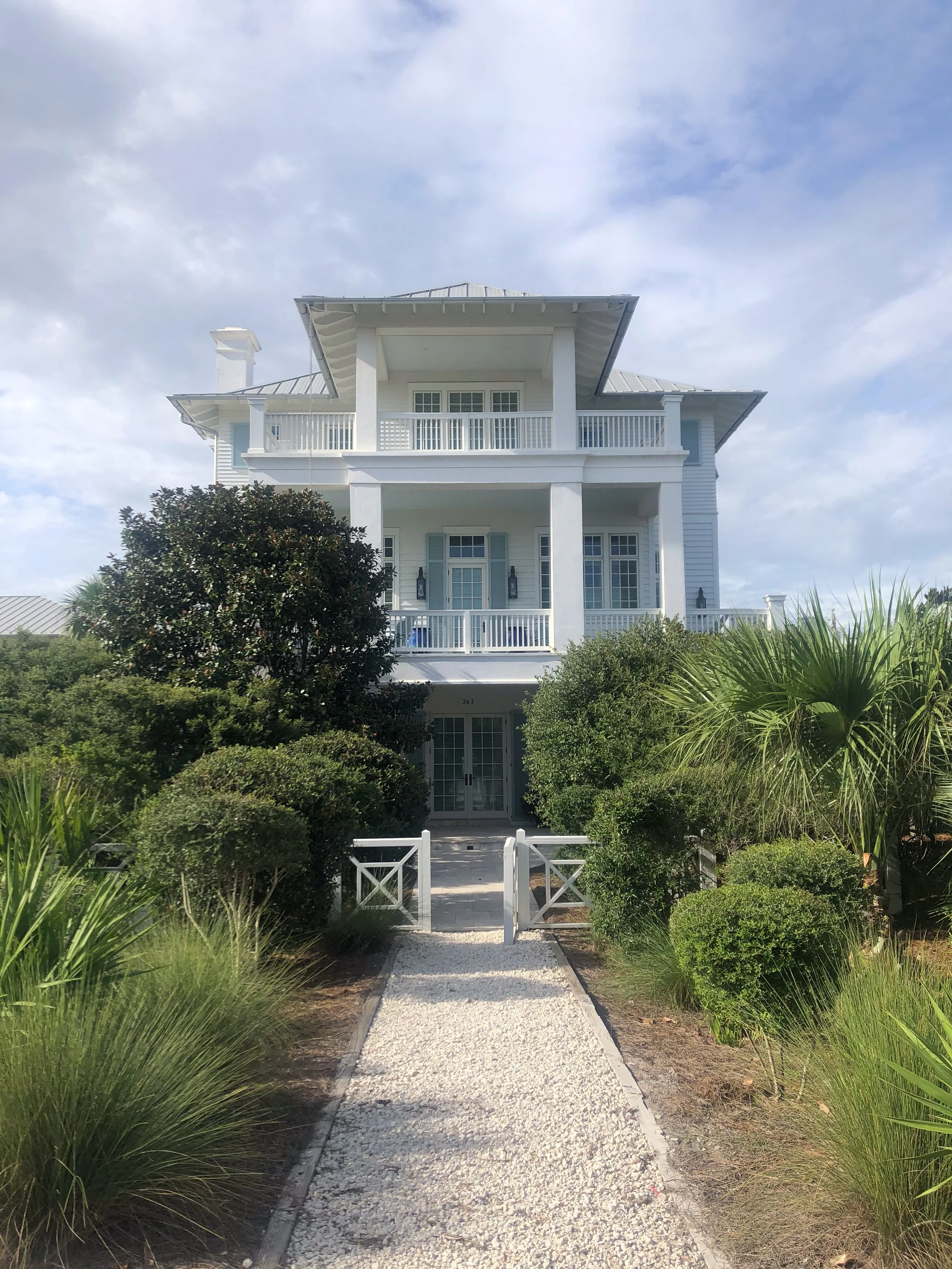 A white multi-story house with a front porch, surrounded by lush green bushes and trees, and a gravel pathway leading to the front gate.